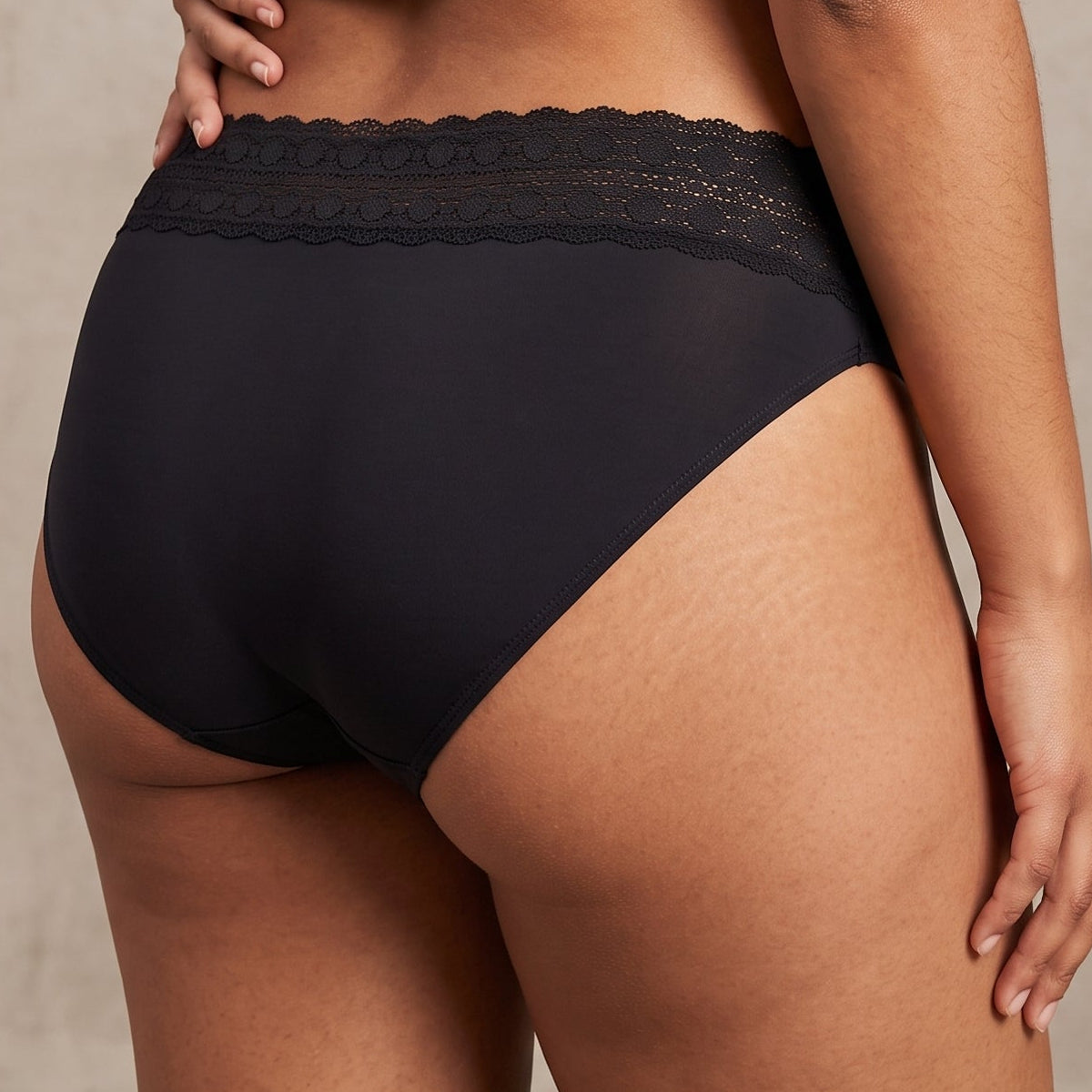 Rear view of a model wearing black high-waisted briefs with a sheer lace waistband, paired with a gray cropped t-shirt and black patterned garter-style stockings, against a textured beige studio background.