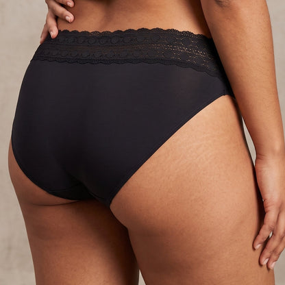 Rear view of a model wearing black high-waisted briefs with a sheer lace waistband, paired with a gray cropped t-shirt and black patterned garter-style stockings, against a textured beige studio background.