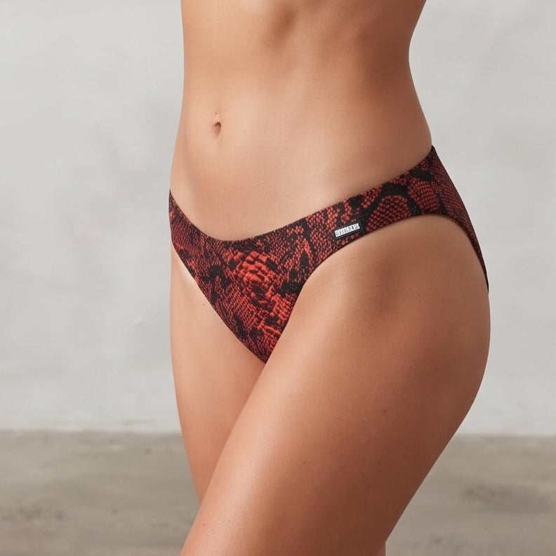 A woman modeling a two-piece bikini with a red and black snake print pattern, standing against a textured light gray wall.