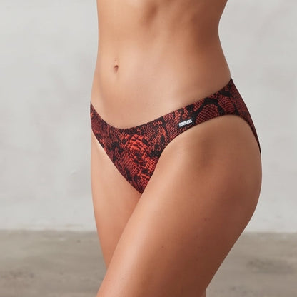 A woman modeling a two-piece bikini with a red and black snake print pattern, standing against a textured light gray wall.
