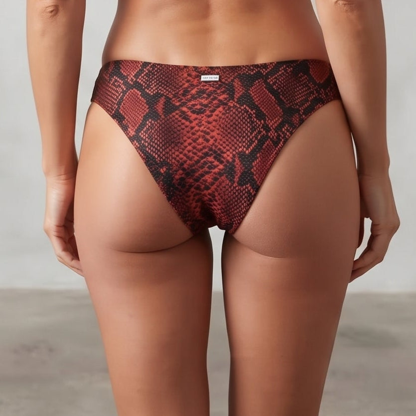 A woman stands with her back to the camera wearing a two-piece bikini with a red and black snakeskin pattern. The bikini features a strappy halter-style top and matching high-cut bottoms.