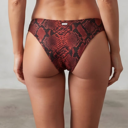 A woman stands with her back to the camera wearing a two-piece bikini with a red and black snakeskin pattern. The bikini features a strappy halter-style top and matching high-cut bottoms.
