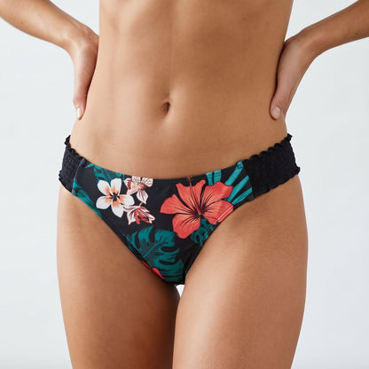 A model wearing black bikini bottoms with a tropical floral print featuring red hibiscus, pink and white flowers, and teal leaves, shown from the waist to upper thighs against a plain light gray studio background.