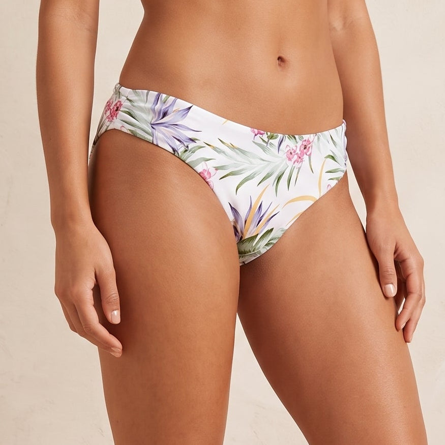 A model wearing a pair of white bikini bottoms with a colorful tropical floral print, standing against a textured beige wall with a woven basket prop.