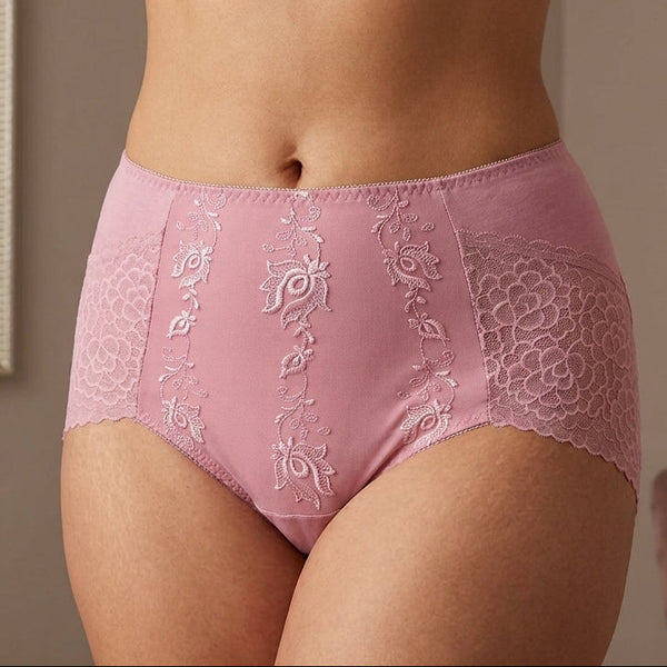 A model wearing high-waisted pink lace panties with embroidered floral details and a matching bra partially visible at the top, photographed indoors against a softly lit neutral background.