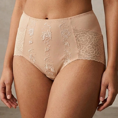 A model wearing high-waisted beige lace panties with embroidered details and a matching bra, shown against a neutral textured studio background.